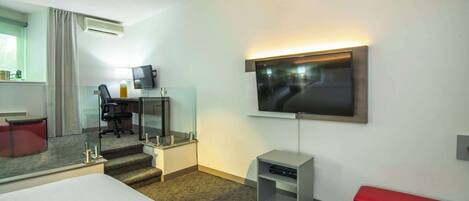 A hotel room with a bed, a desk with a chair, a TV mounted on the wall, and a small bench.