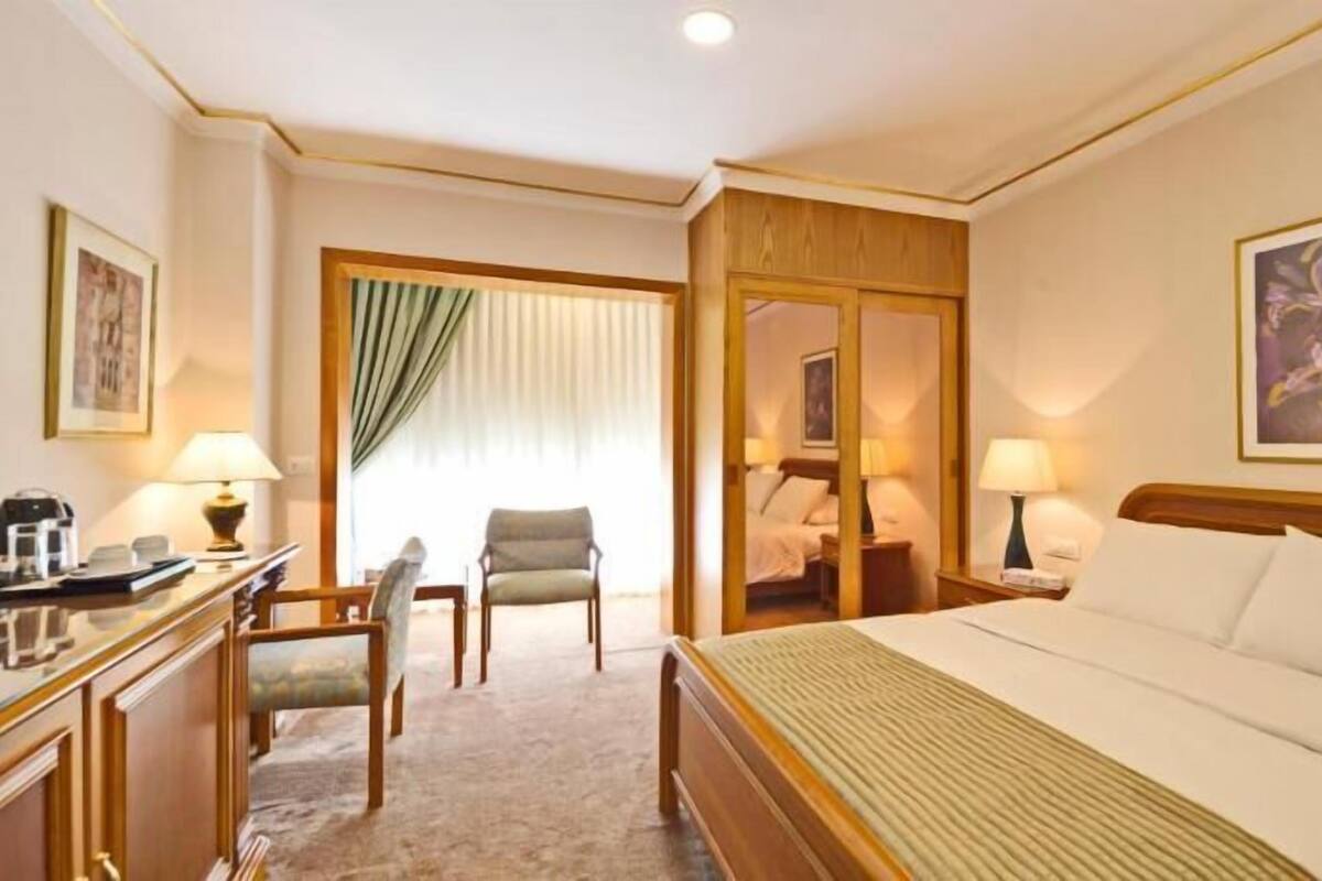 A hotel room with a bed, desk, chair, and a view of another room through a door.