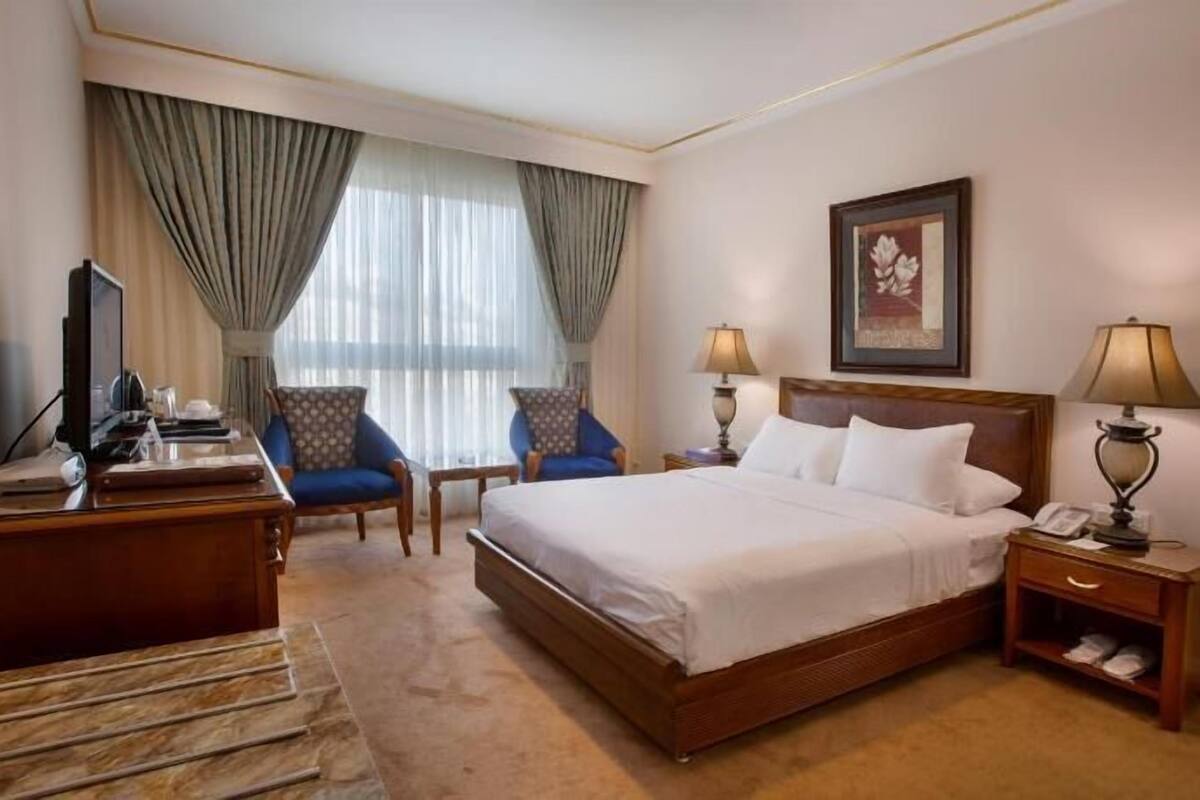 A hotel room with a bed, two chairs, a desk with a computer, a lamp, and a painting on the wall.
