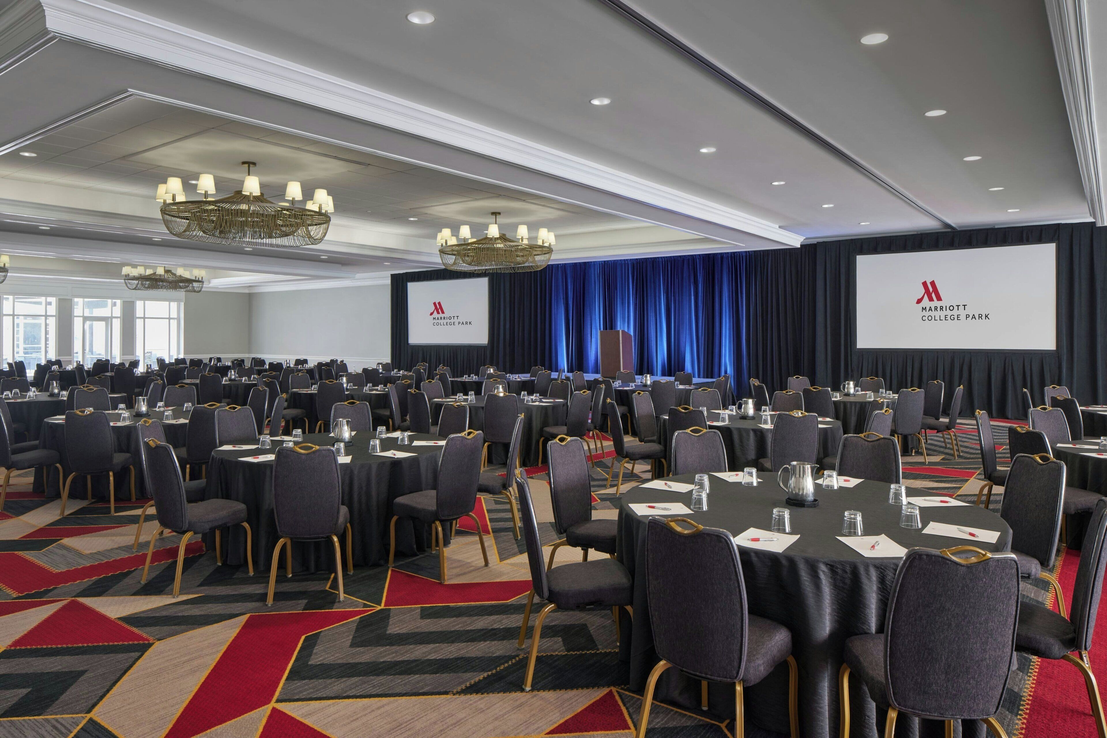 Un salón de conferencias con mesas redondas, sillas y una pantalla grande mostrando el logotipo del Marriott College Park.