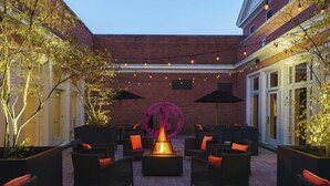 An outdoor seating area with wicker furniture and a central fire feature, surrounded by brick walls and string lights. - College Park Marriott Hotel & Conference Center (College Park)