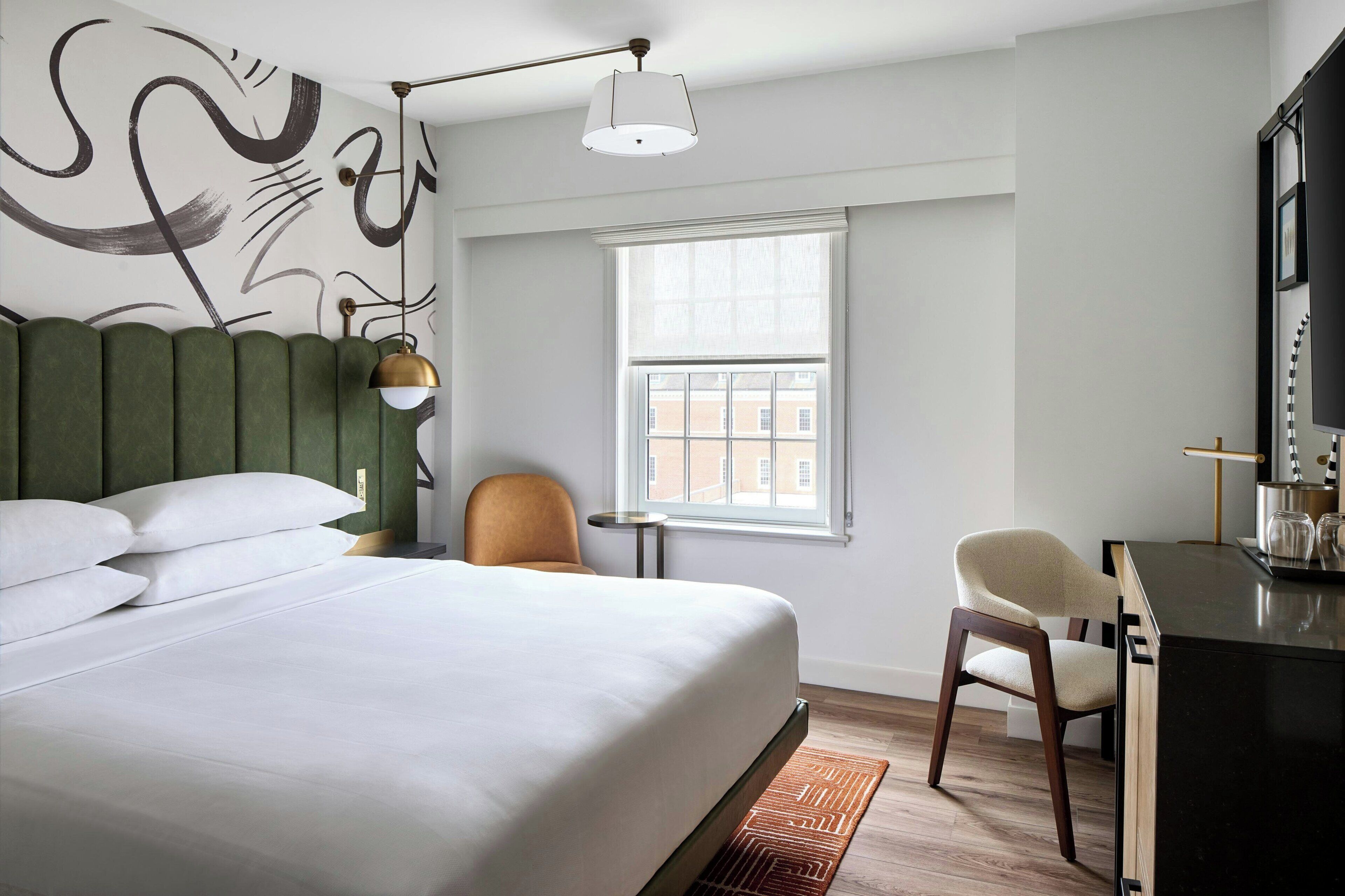 A hotel room with a bed, a desk, a chair, a window, and a wall with abstract art.