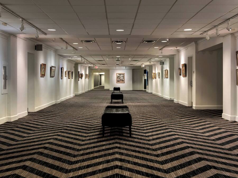 A long hallway with a patterned carpet, framed pictures on the walls, and a bench in the center.