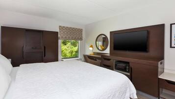 A hotel room with a large bed, a desk with a flat-screen TV, a mirror, and a window with a view of greenery.