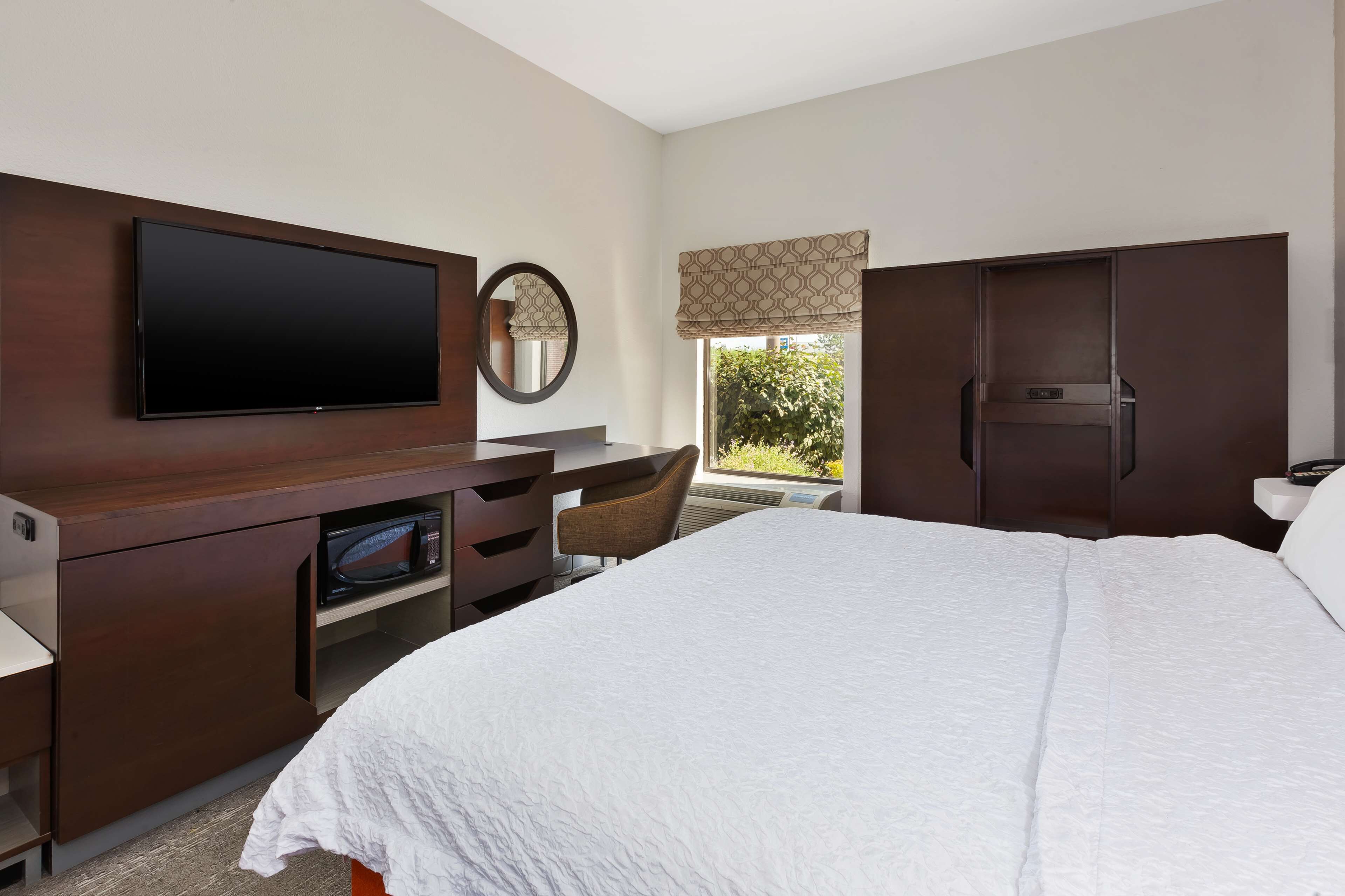 Quarto de hotel com uma cama grande, televisão de tela plana, escrivaninha com micro-ondas, uma cadeira e guarda-roupa.