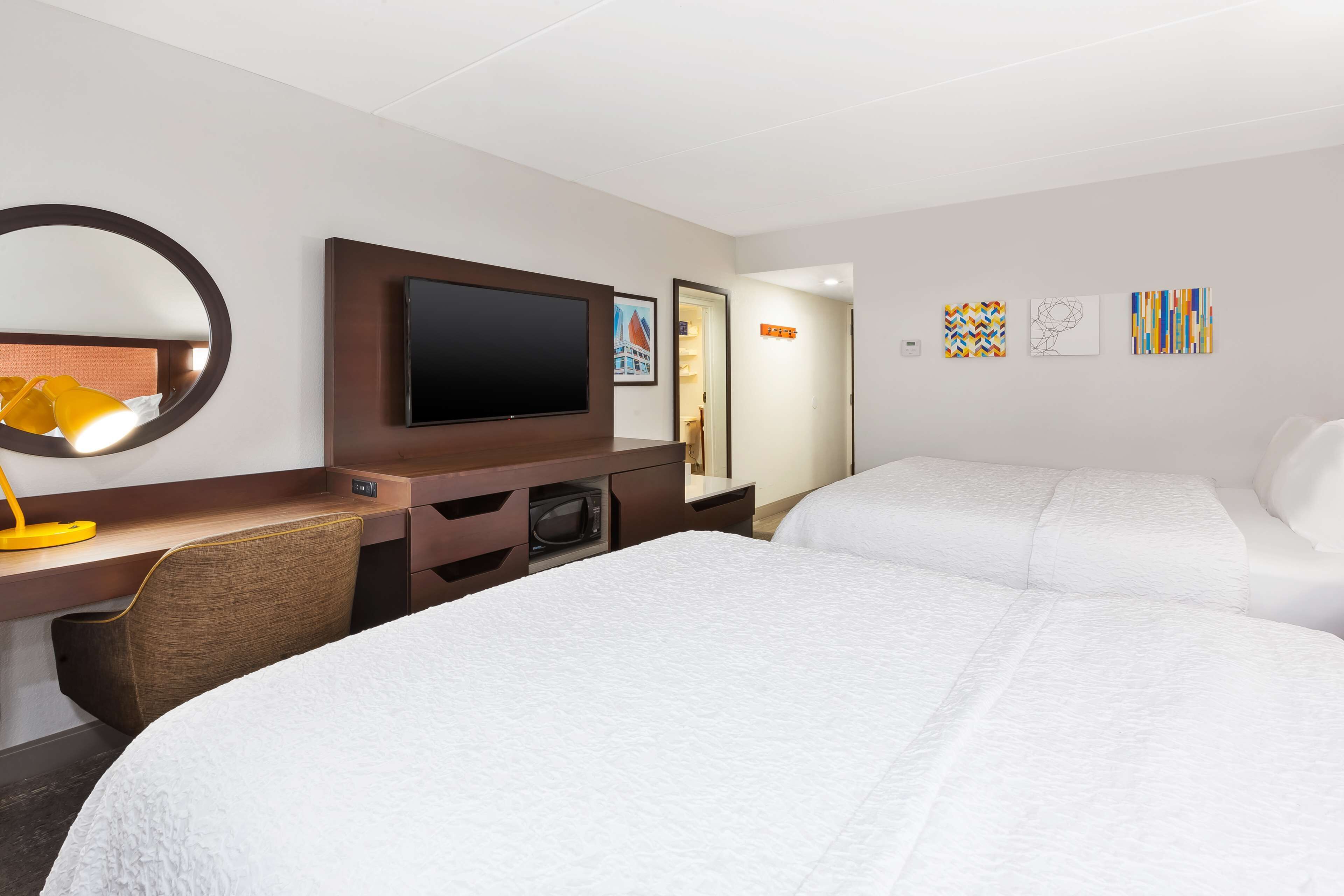 Habitación de hotel con una cama, un escritorio con televisor, un espejo y obras de arte en la pared.