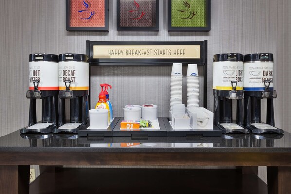 A breakfast bar with coffee dispensers labeled "Hot Water," "Decaf Roast," and "Light Roast," along with stacks of white cups and a "Happy Breakfast Starts Here" sign.