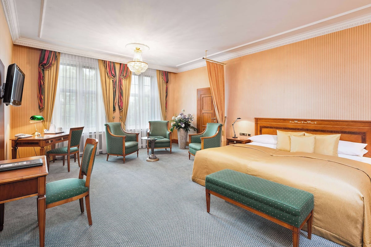 A hotel room with a large bed, two armchairs, a desk with a lamp, a television, and a window with curtains.