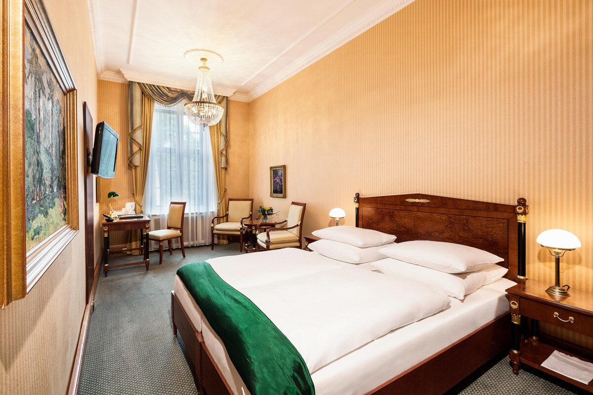 Une chambre d’hôtel avec un grand lit, deux fauteuils, un bureau, une télévision et un lustre.
