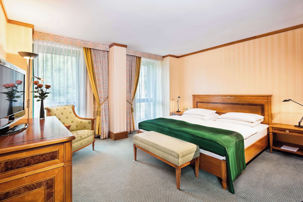A hotel room with a bed, a desk with a TV, a chair, a bedside table, and a lamp.