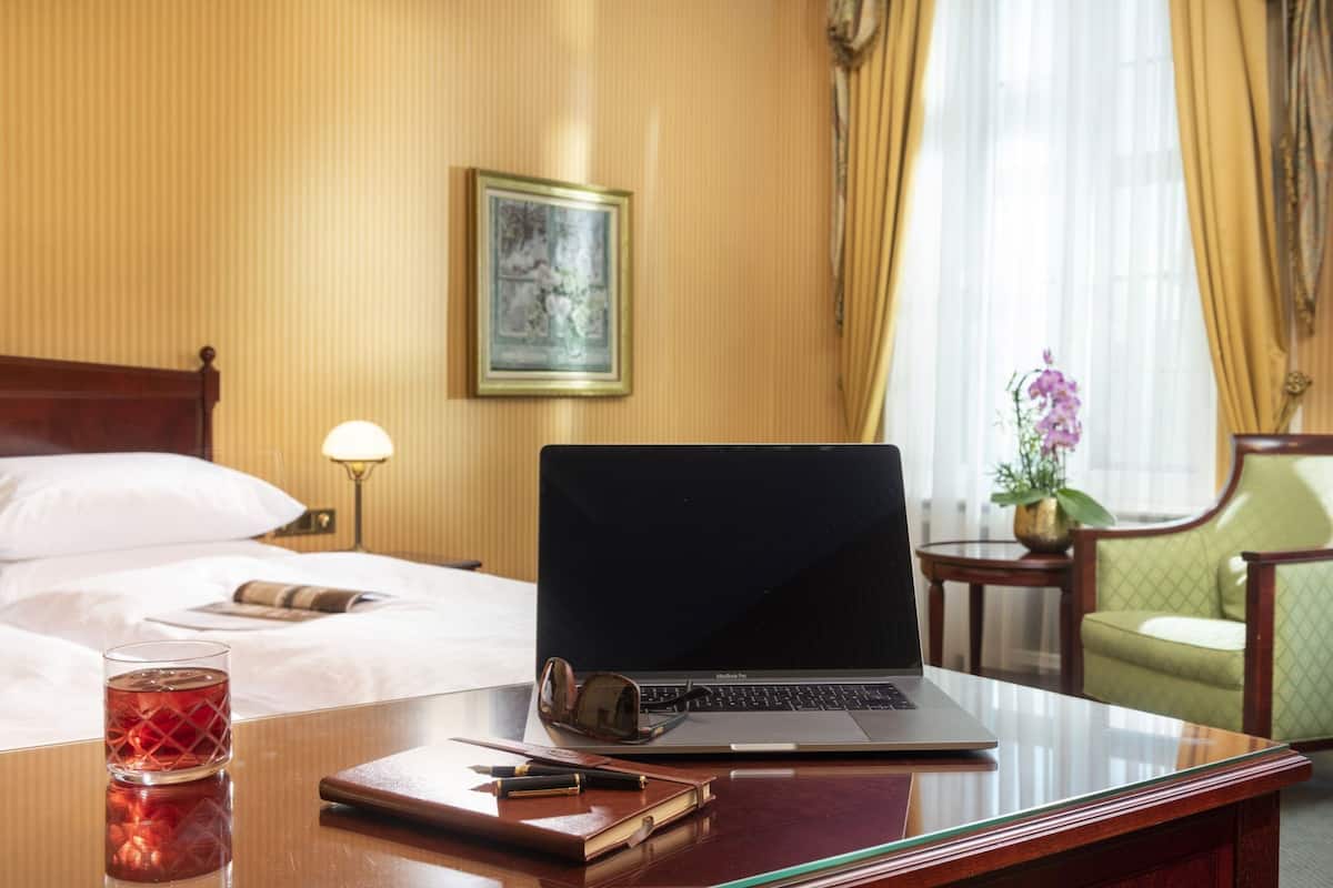 A hotel room with a bed, a desk with a laptop, a lamp, a chair, and a vase with flowers.
