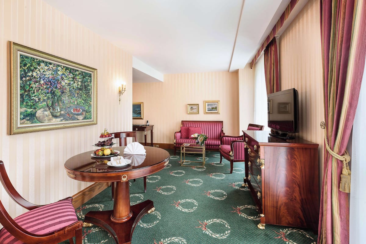 A hotel room with a round dining table, two chairs, a sofa, a TV, and a decorative painting.