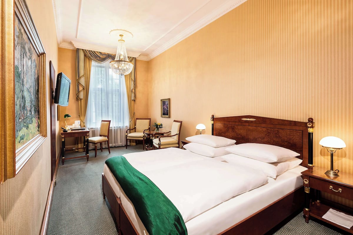 A hotel room with a large bed, a desk with a chair, a television, and a chandelier.