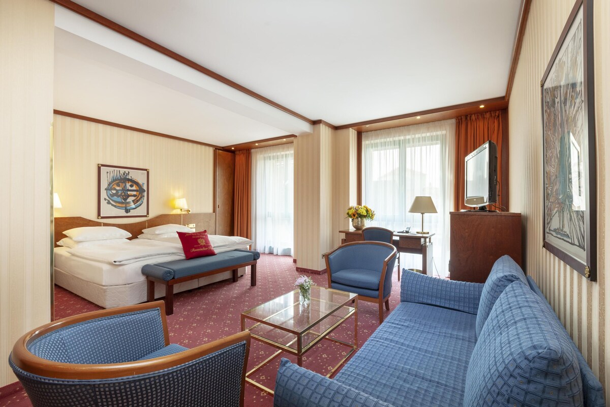 A hotel room with a bed, a sofa, a chair, a coffee table, a desk with a lamp, and a TV.