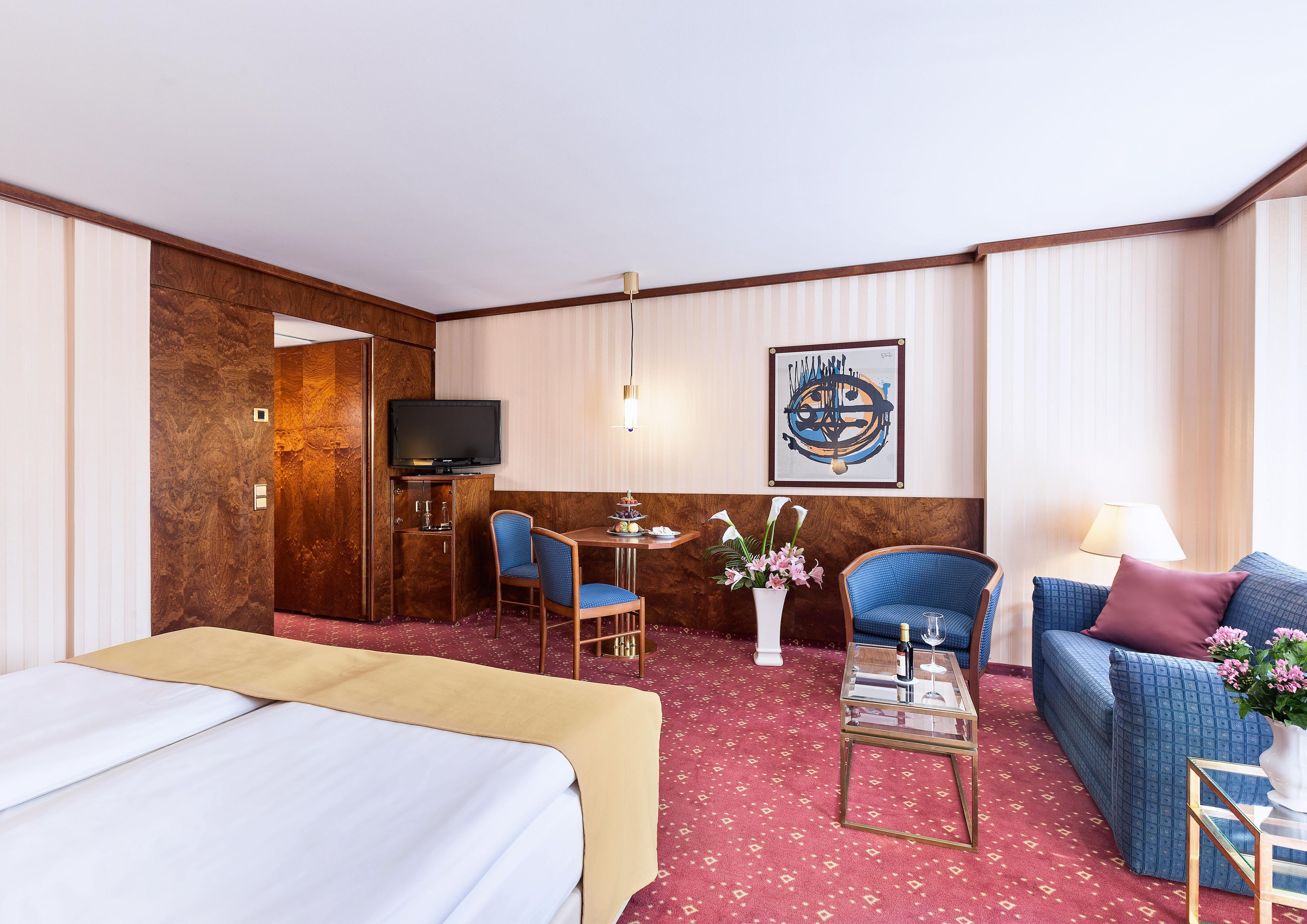 A hotel room with a bed, a desk with a television, a seating area with a sofa and chairs, and a decorative wall piece.