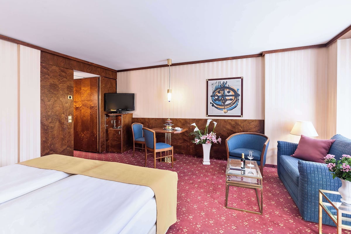 A hotel room with a bed, a sofa, a chair, a small table with a vase of flowers, a television, and a wall-mounted artwork.
