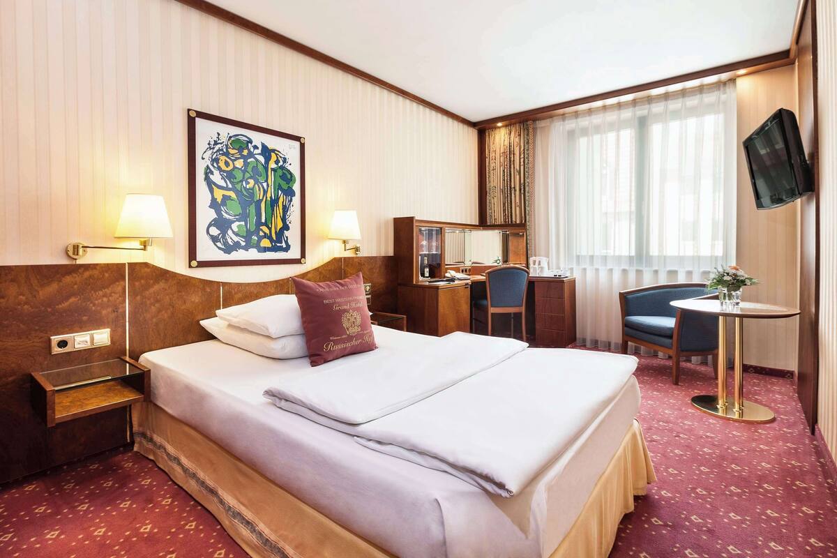 A hotel room with a large bed, a desk, a chair, a TV, and artwork on the wall.