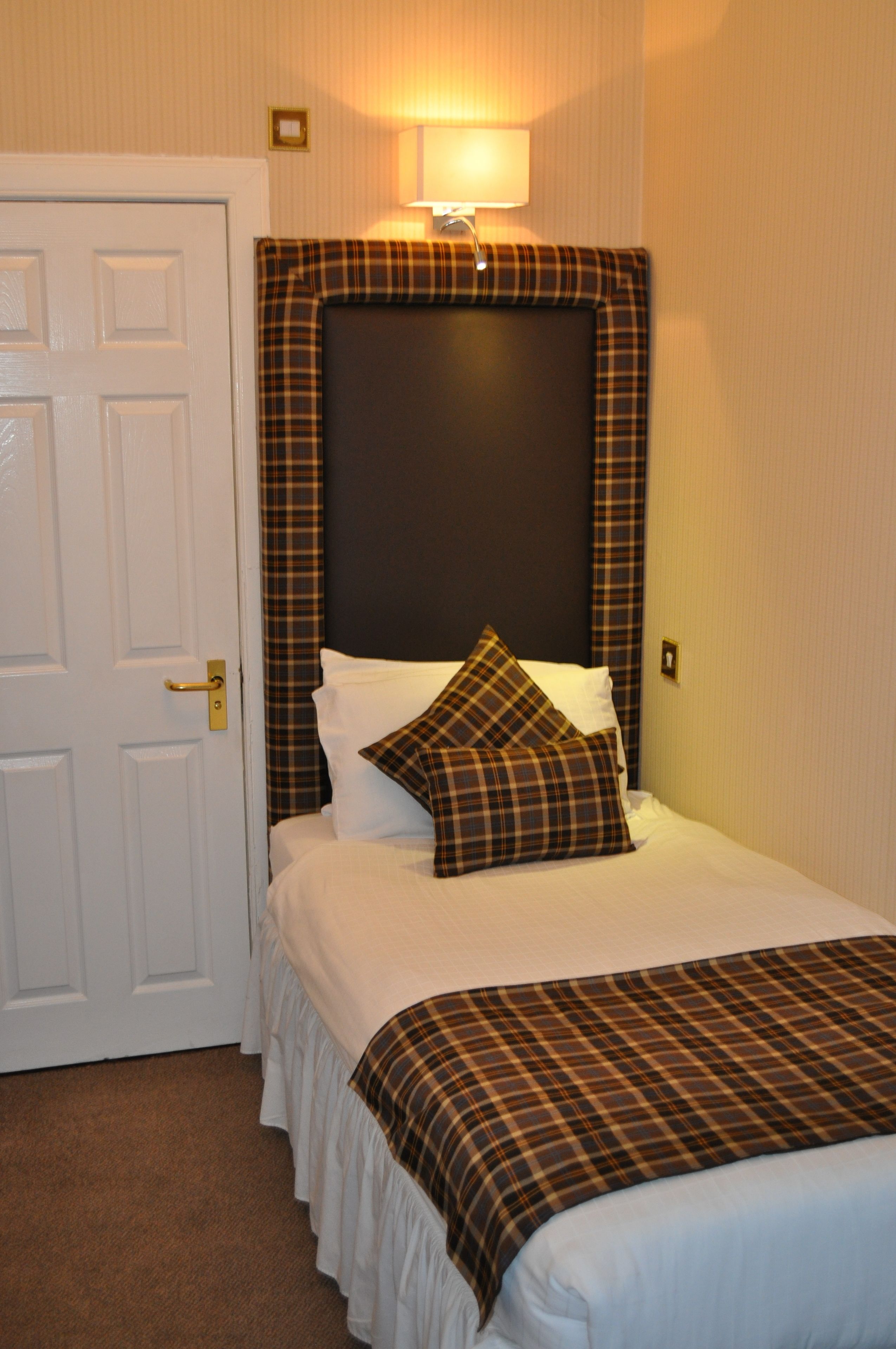 A neatly made bed with a plaid headboard, a bedside lamp, and a white door.