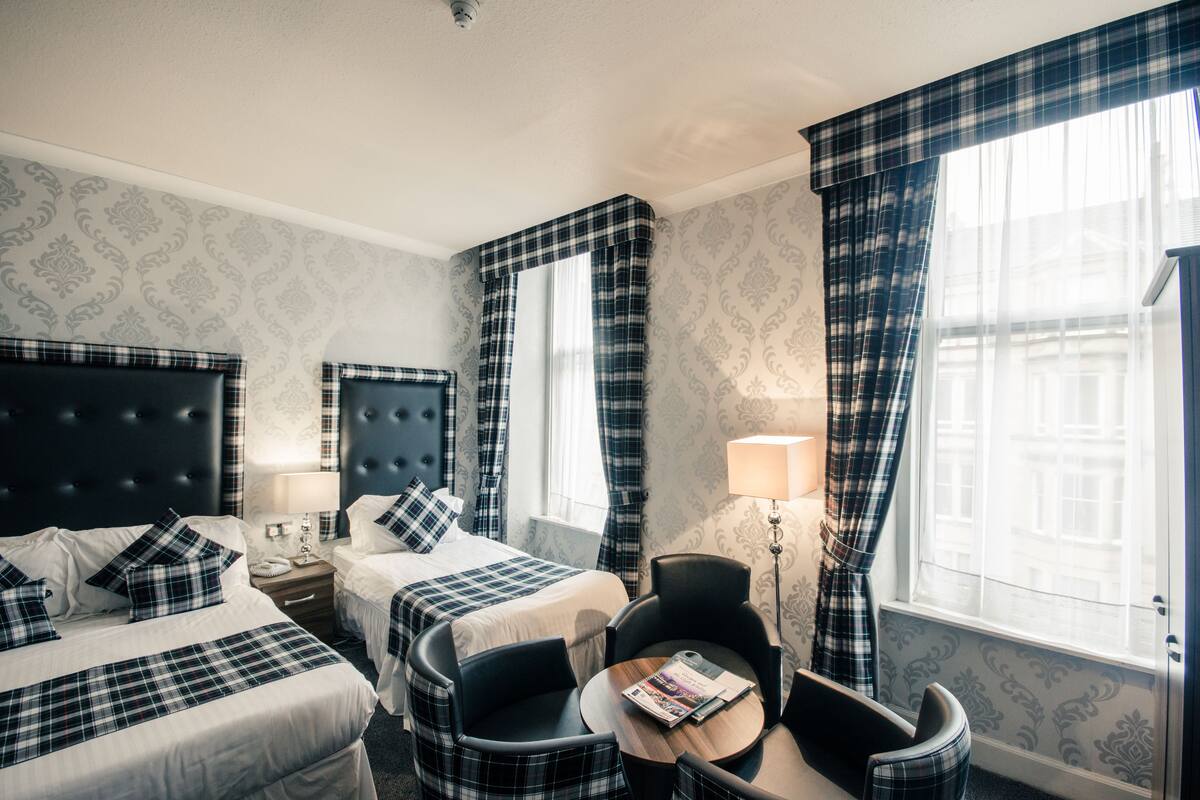 A hotel room with two beds, a desk, a chair, a table, and a lamp.