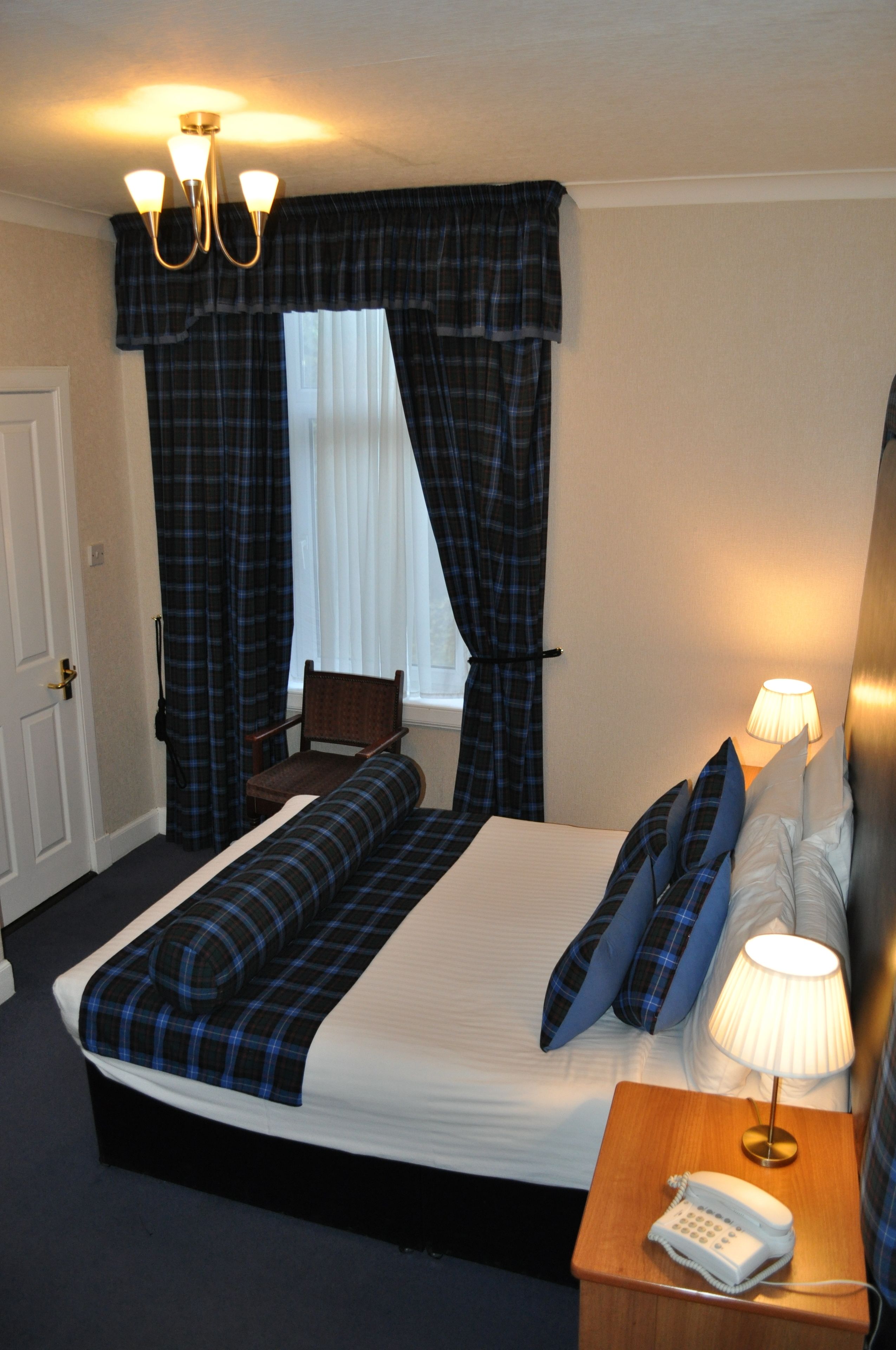 A hotel room with a large bed, a bedside table with a lamp, a chair, and a window with curtains.