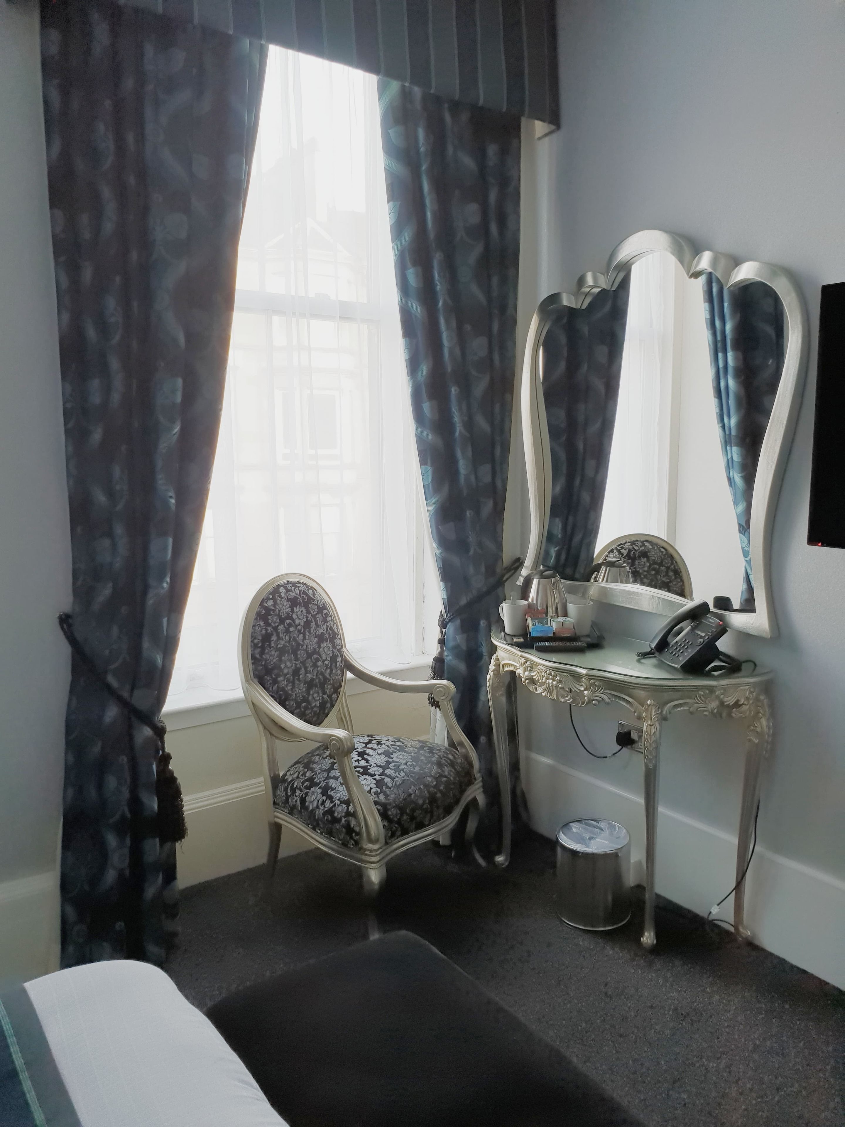 Een hotelkamer met een bed, een stoel, een kaptafel met spiegel, een telefoon en een raam met gordijnen.