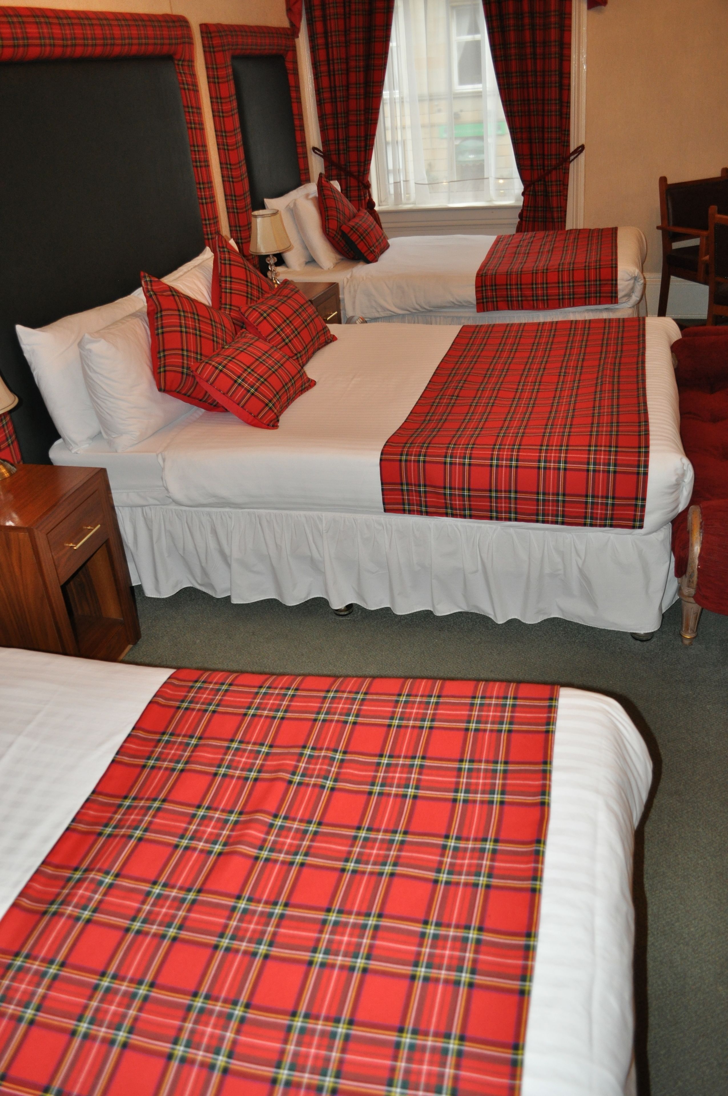 A hotel room with two beds, each with a red plaid bedspread and white bedding. There is a wooden nightstand with a lamp, and a chair in the corner.