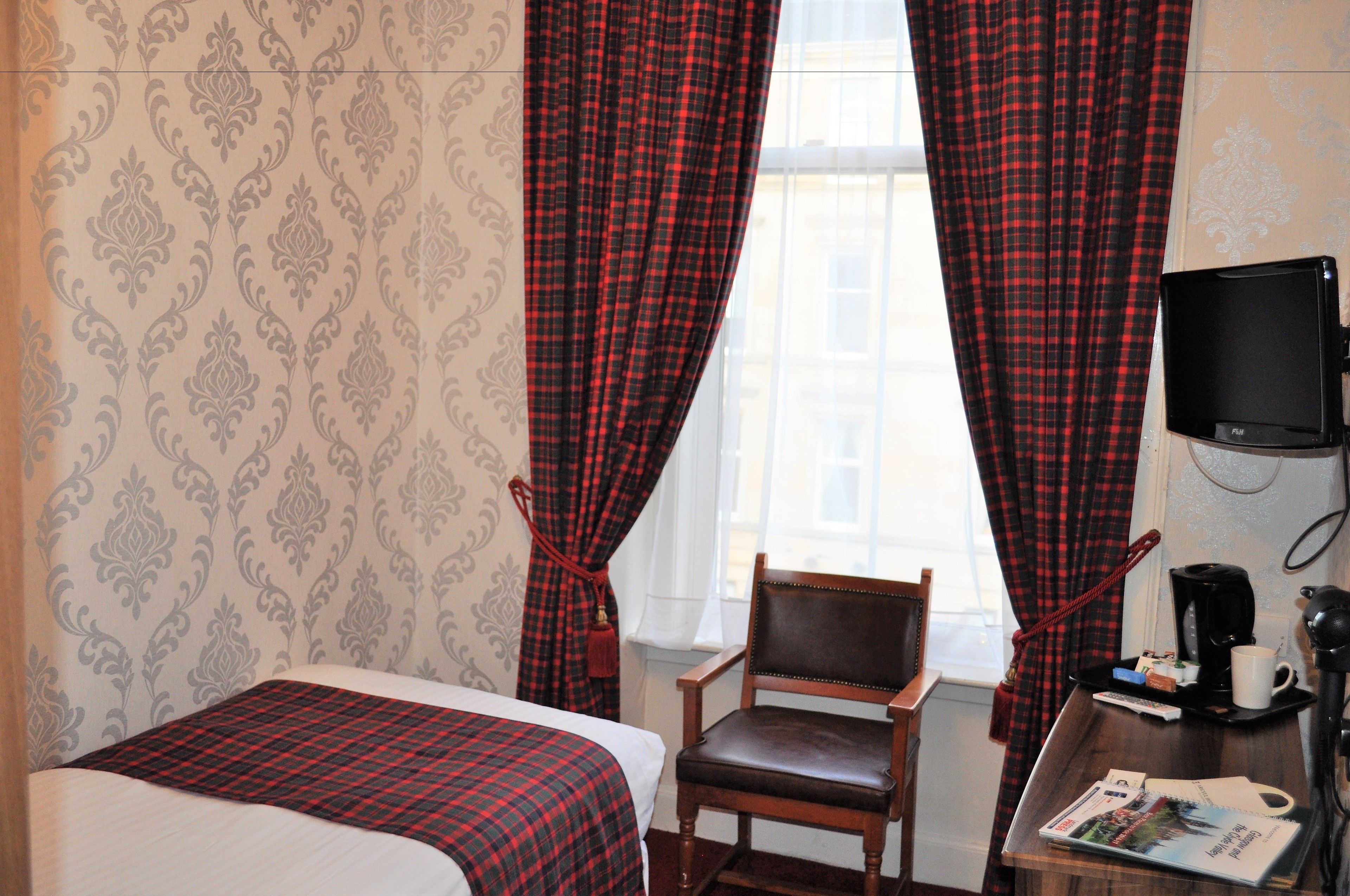 A hotel room with a bed, a chair, a desk with a coffee maker, and a TV on the wall.