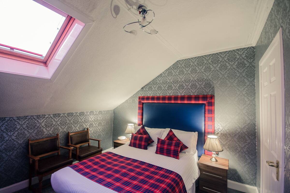 A bedroom with a skylight, a bed with a plaid bedspread, two nightstands with lamps, and a headboard with a red and blue design.