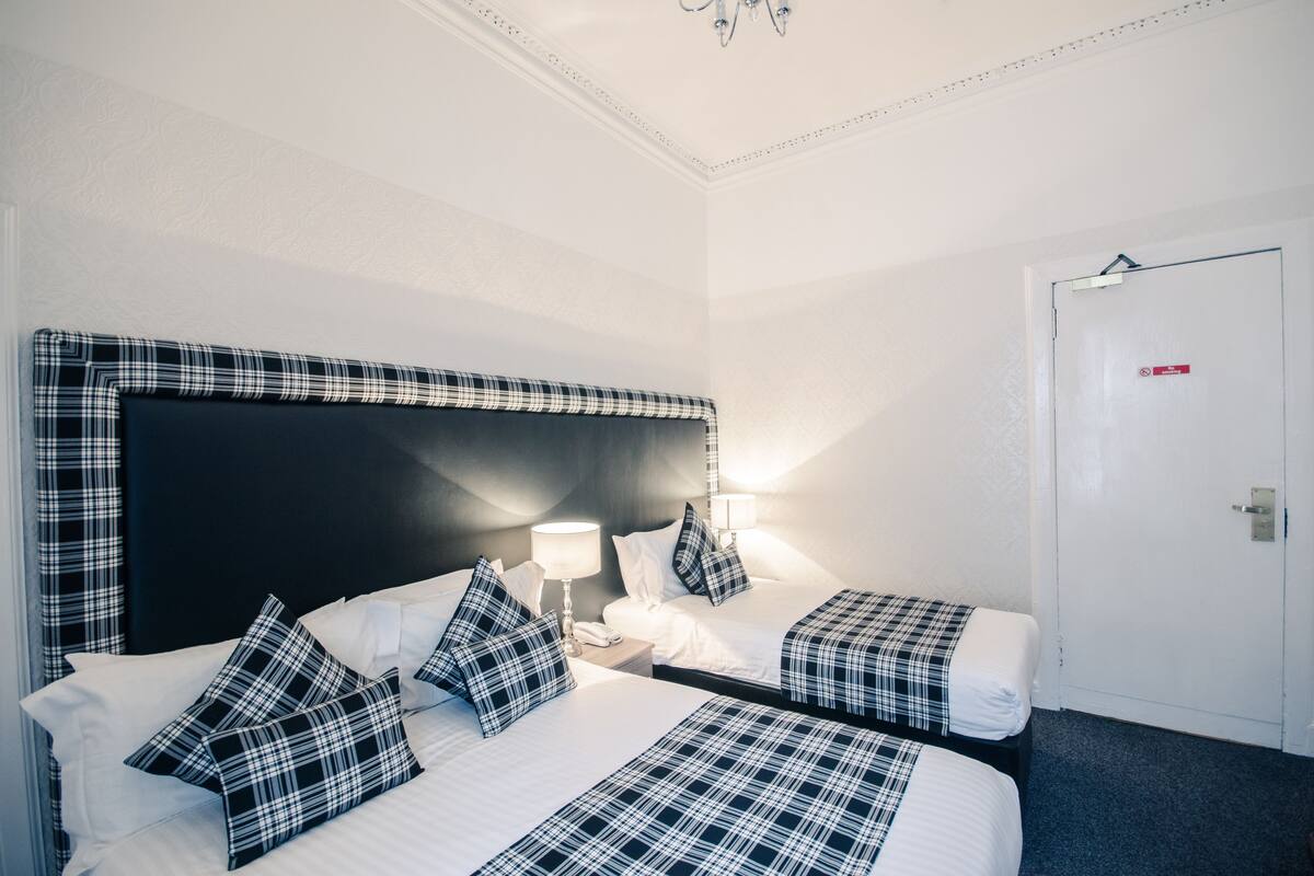 A hotel room with two beds, a black headboard, white walls, a door with a red sign, and a chandelier.