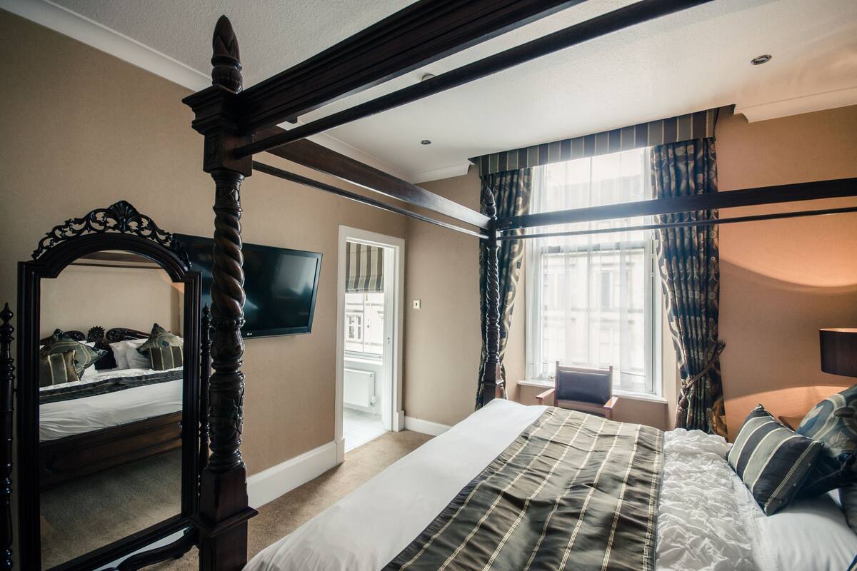 A bedroom with a four-poster bed, a large mirror, a television, and a window with curtains.