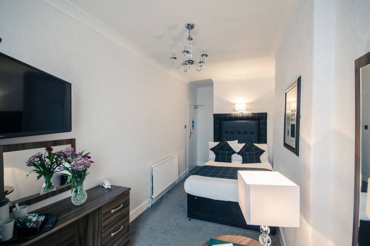 A hotel room with a large flat-screen TV, a bed with a dark headboard, a bedside table with a lamp, and a dresser with a mirror.