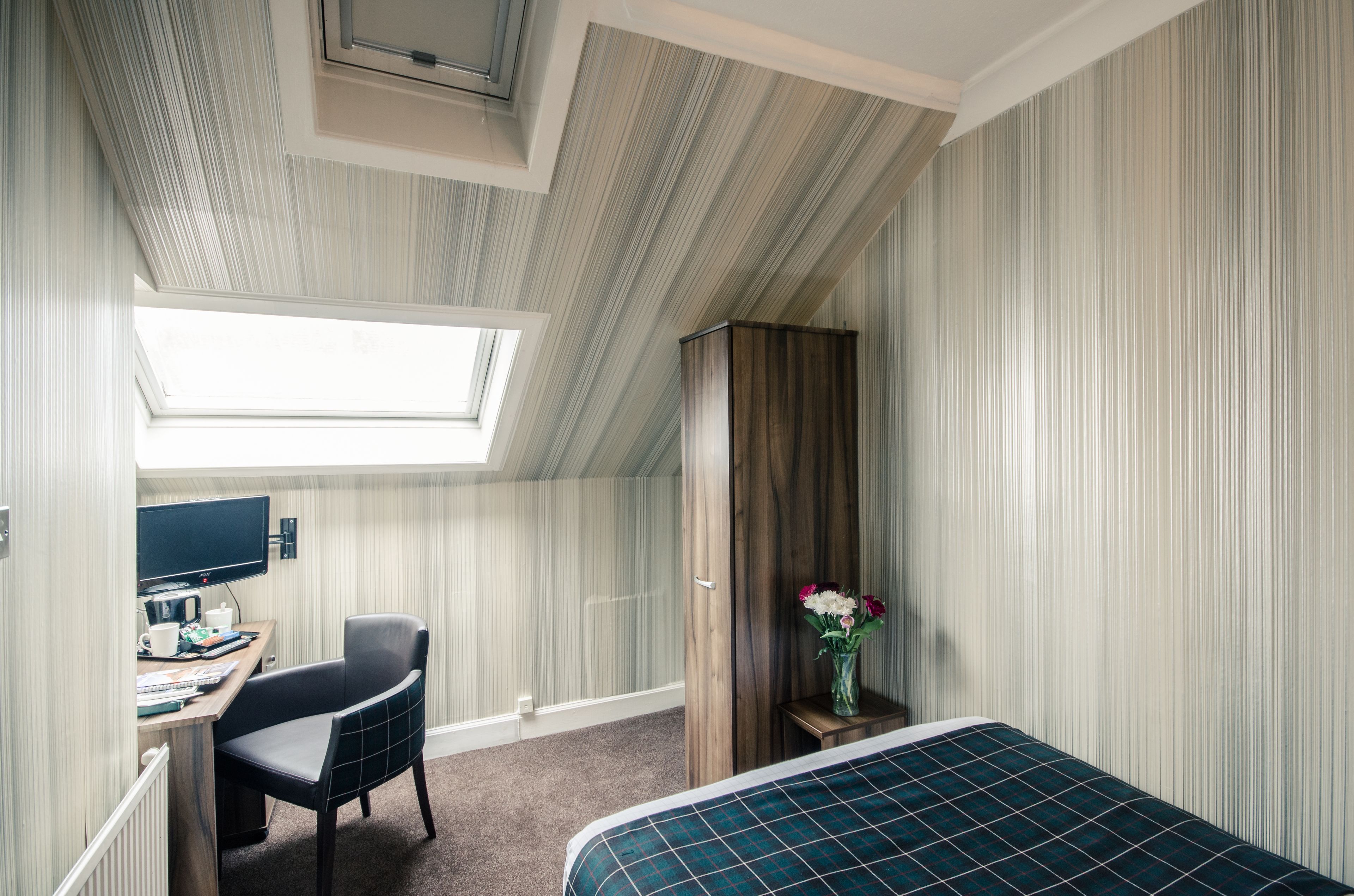 Ein Hotelzimmer mit schräg stehender Decke, einem Schreibtisch mit Stuhl, einem Bett und einem kleinen Tisch mit Blumen.