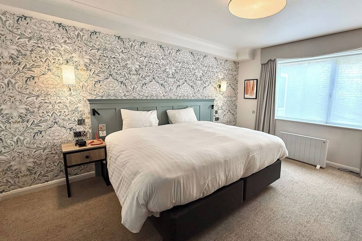 Ein Hotelzimmer mit einem großen Bett, einem Nachttisch, einem Sessel, einem Fenster mit Vorhängen und einer Wand mit Muster.