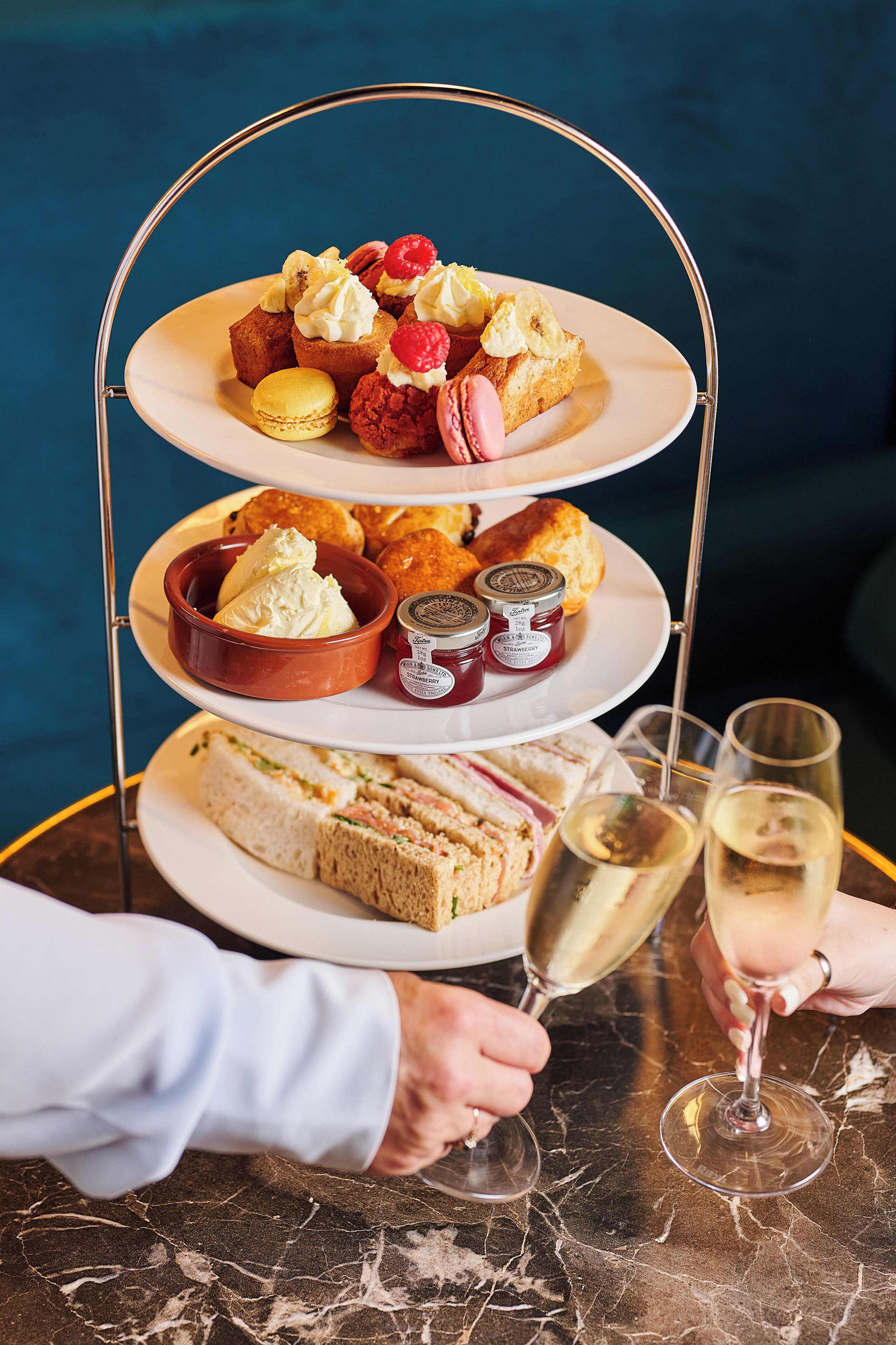A tiered dessert stand with macarons, pastries, and sandwiches, accompanied by two glasses of champagne.