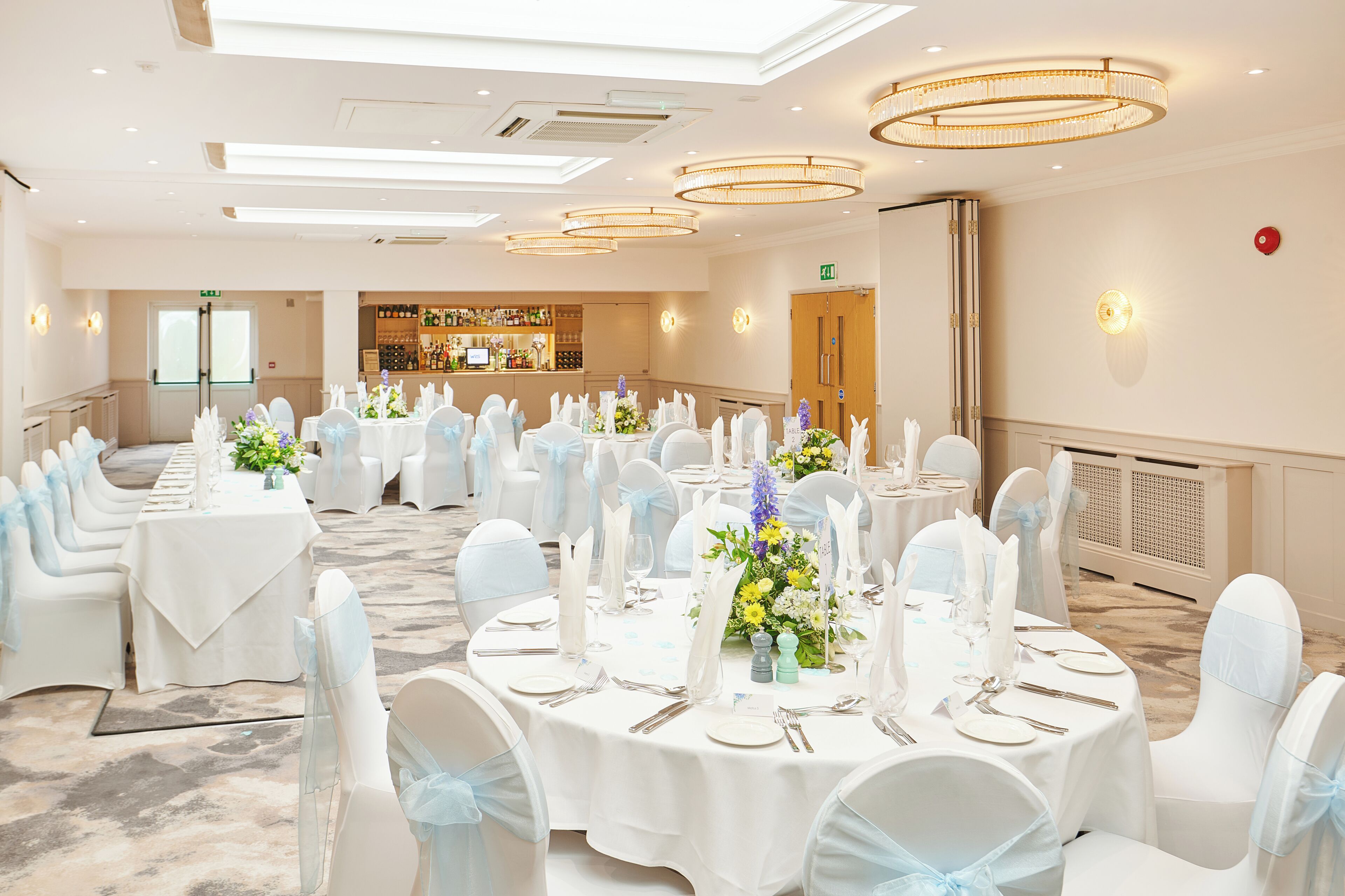Un salón de banquetes con mesas redondas dispuestas para un evento formal, destacando manteles blancos, sillas con cintas azules y centros de mesa florales.