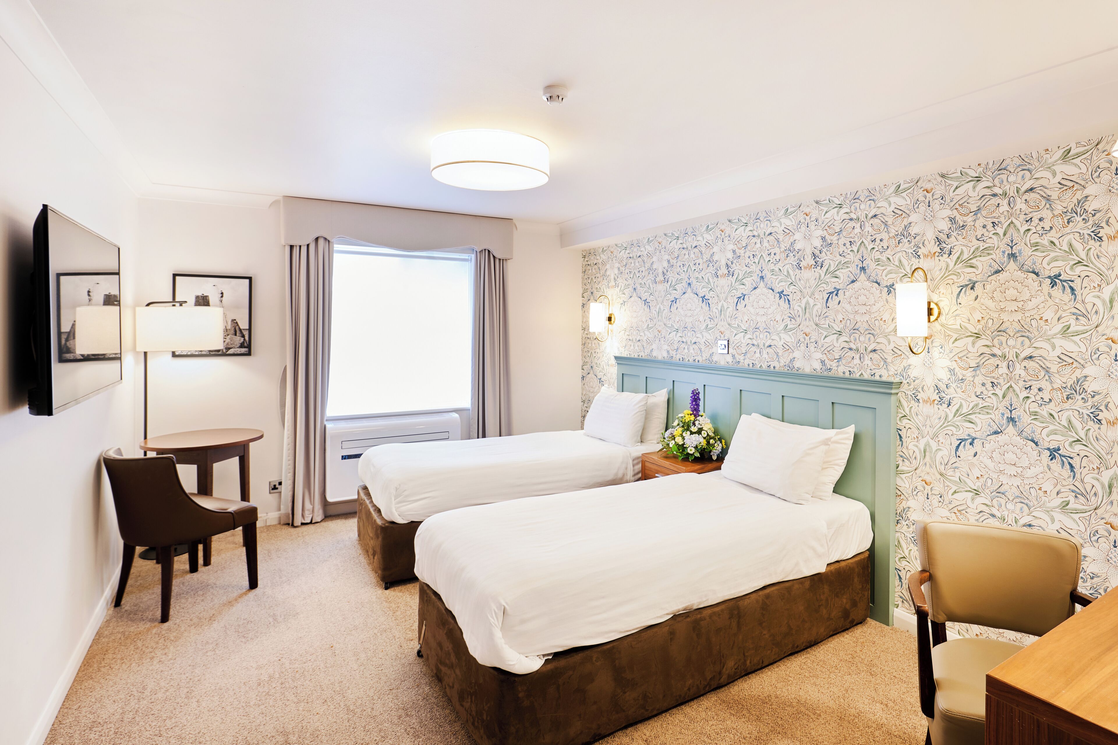 A hotel room with two beds, a desk, a chair, a lamp, and a window.