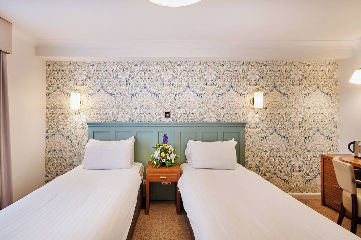 Ein Hotelzimmer mit zwei Betten, einem Nachttisch aus Holz mit Blumenvase und einem Schreibtisch mit Stuhl.