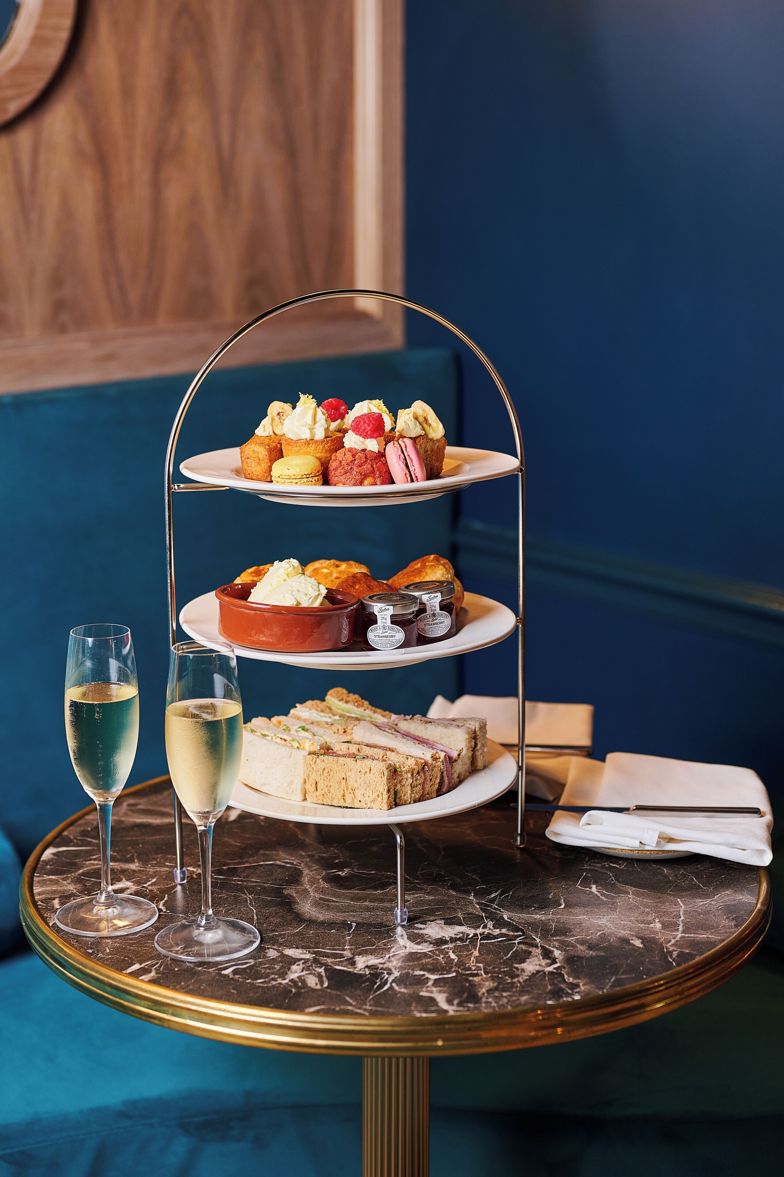 A three-tiered dessert stand with various pastries and sandwiches on a marble table, accompanied by two glasses of champagne.