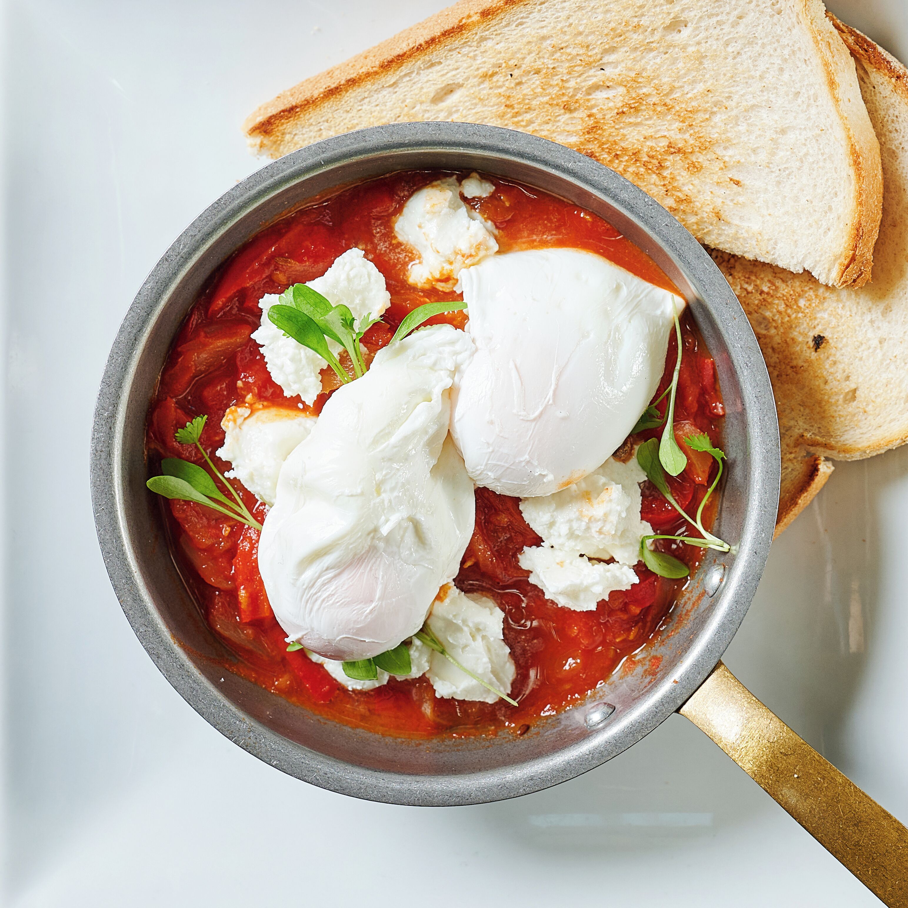 Uma tigela de sopa de tomate com ovos pochê e vegetais frescos, acompanhada de fatias de pão torrado.