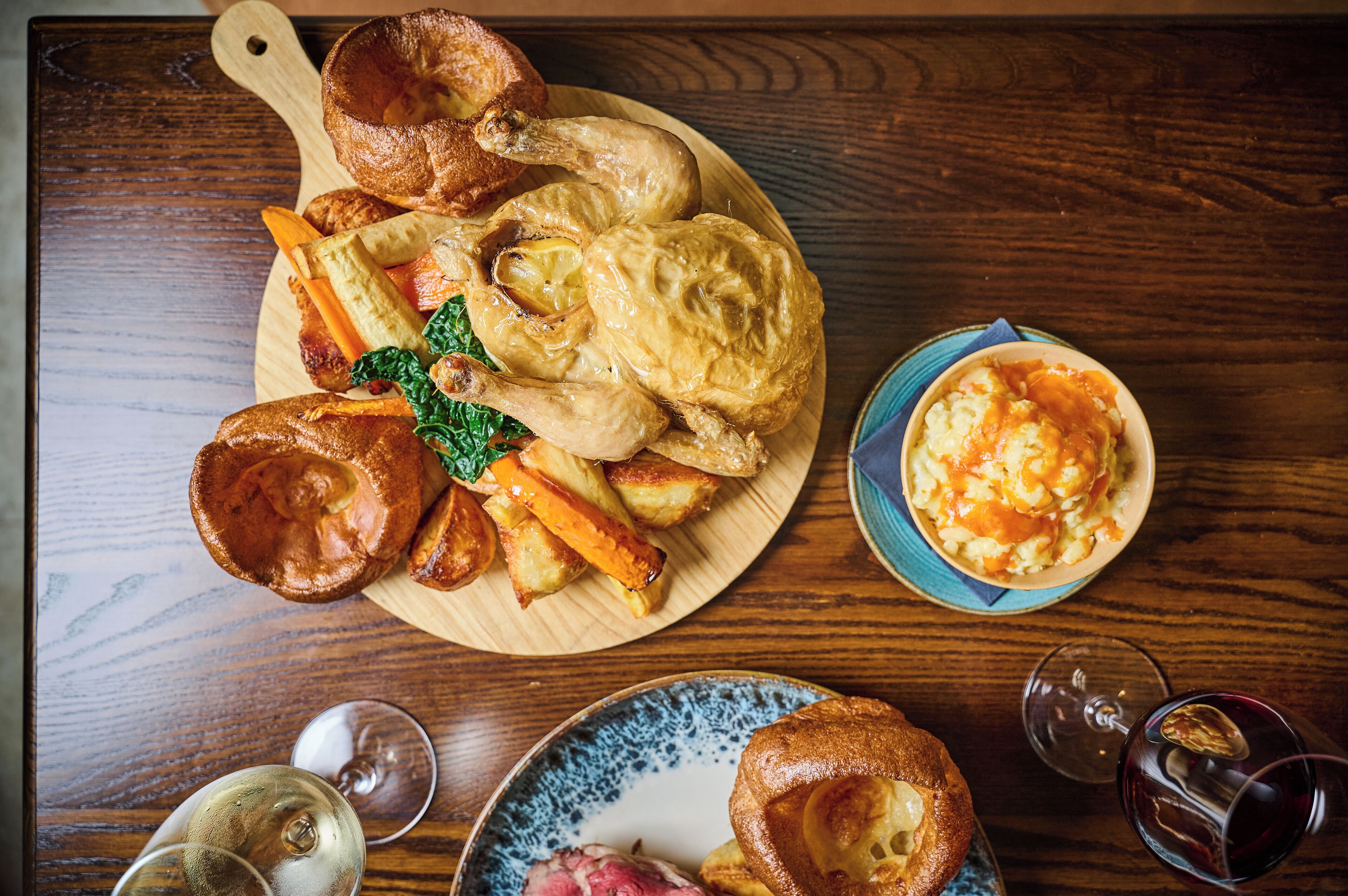 Een houten tafel met een gebraden kip, Yorkshire pudding, geroosterde aardappelen en een schaal met gepureerde aardappelen.