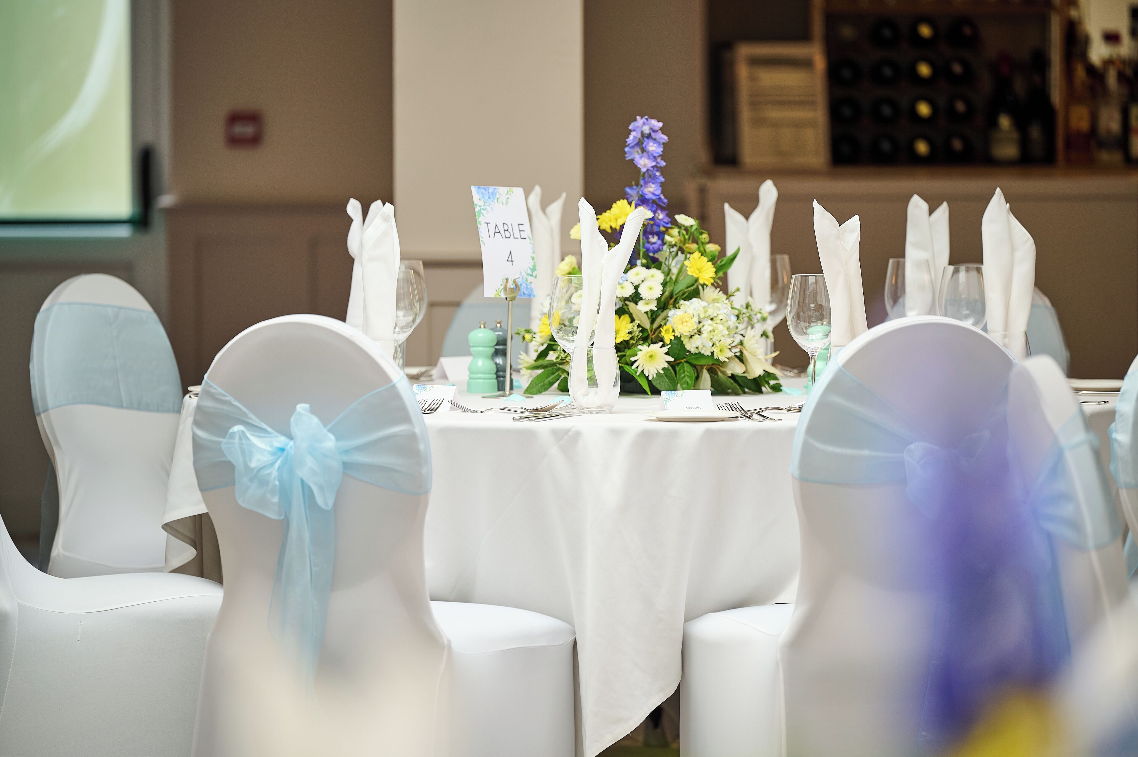 A table set for a formal event with white tablecloths, white chairs with blue sashes, and a floral centerpiece.