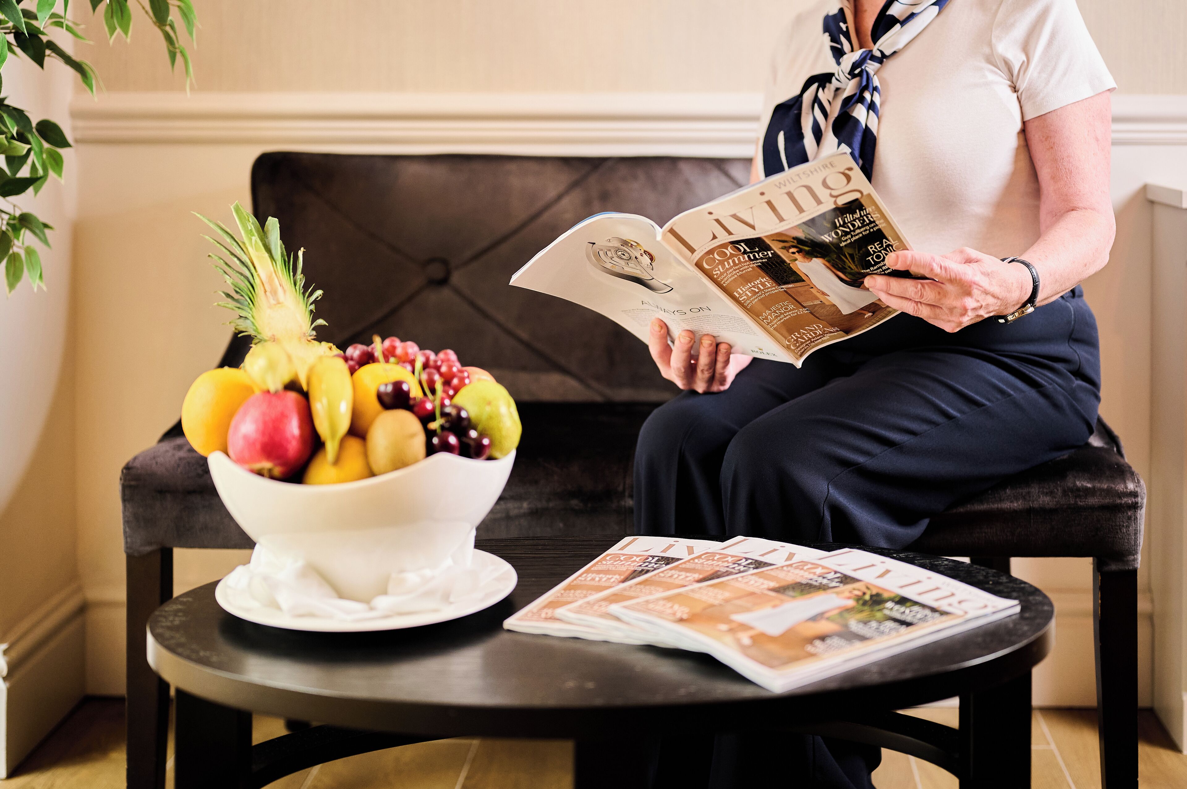 A person reading a magazine with a bowl of fruit on a table.