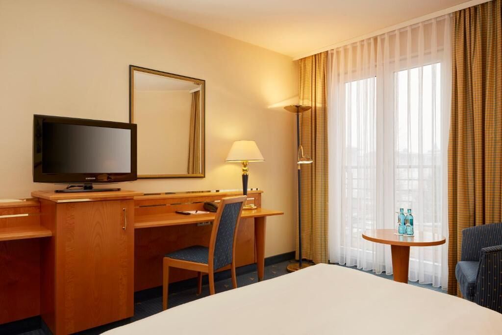 A hotel room with a bed, a desk with a television, a chair, a lamp, a small table with bottles, and large windows with curtains.