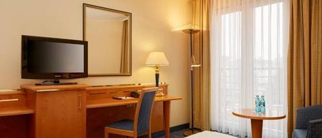 A hotel room with a bed, a desk with a television, a chair, a lamp, a small table with bottles, and large windows with curtains.