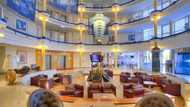 A spacious lobby with multiple seating areas, a central decorative piece, and a large screen. - ACHAT Hotel Magdeburg (Magdeburg)