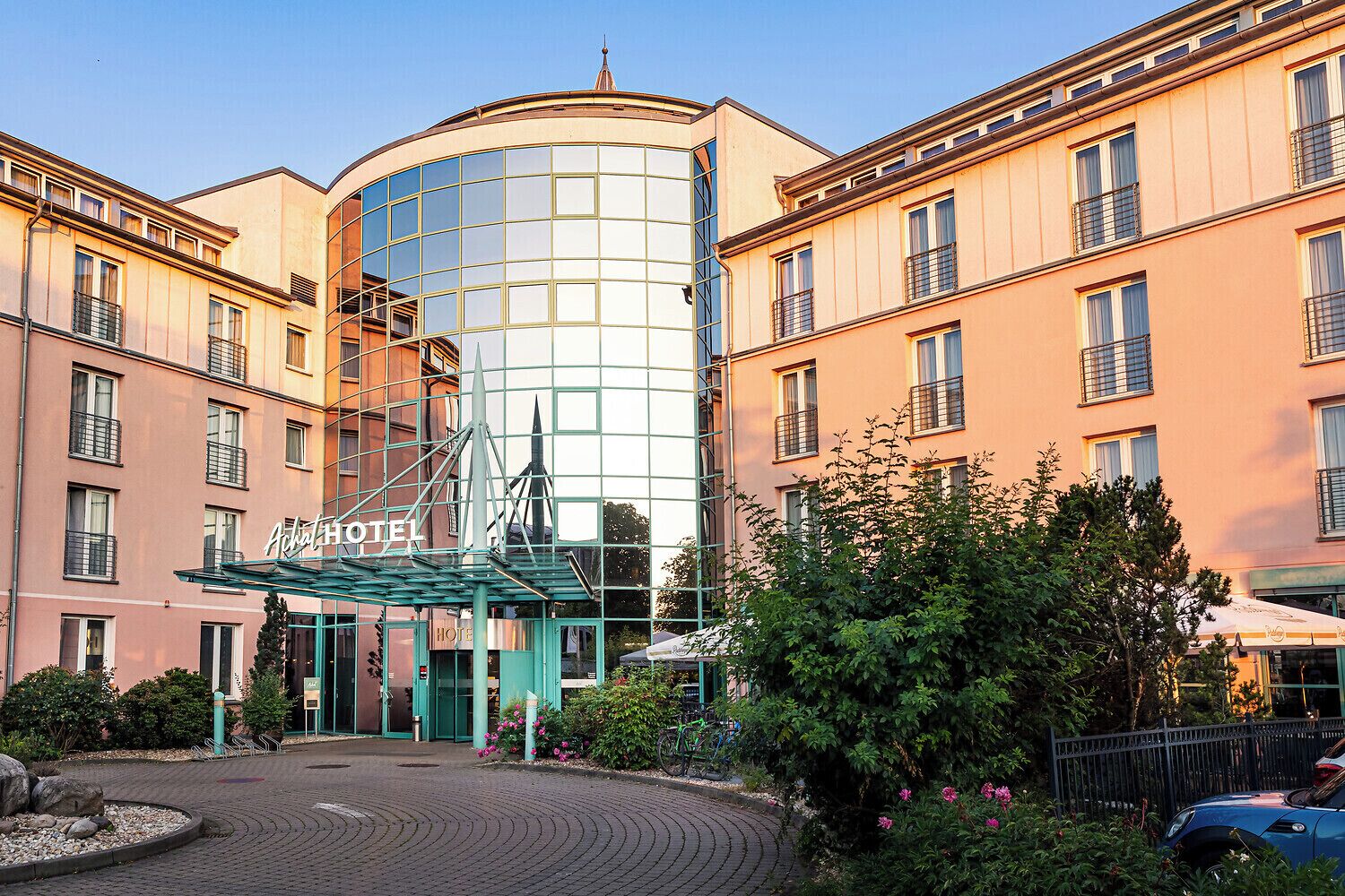 Un hotel con fachada de vidrio moderno, entrada central con toldo verde y zonas ajardinadas circundantes.