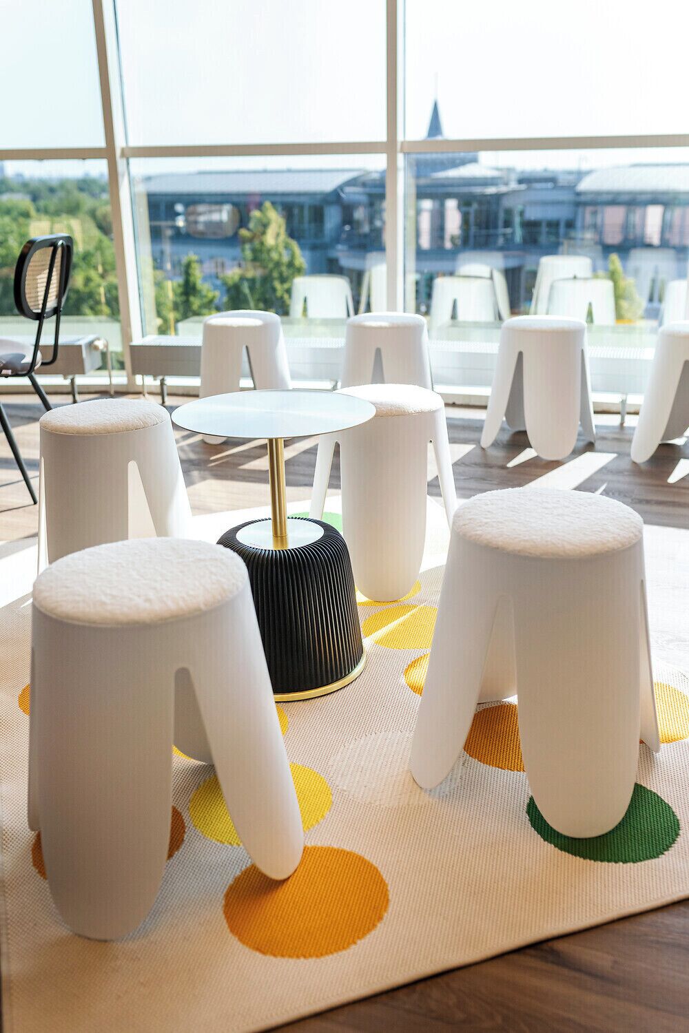 A room with a round table and white stools, a patterned mat, and a view of buildings through large windows.