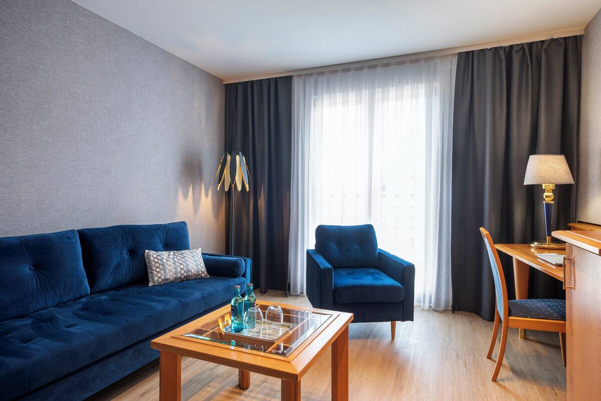 A modern hotel room with a blue sofa, a wooden coffee table, a blue armchair, and a desk with a lamp.