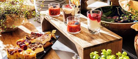 Una mesa de madera con diversos alimentos y bebidas, rodeada de plantas en macetas y una pila de platos.