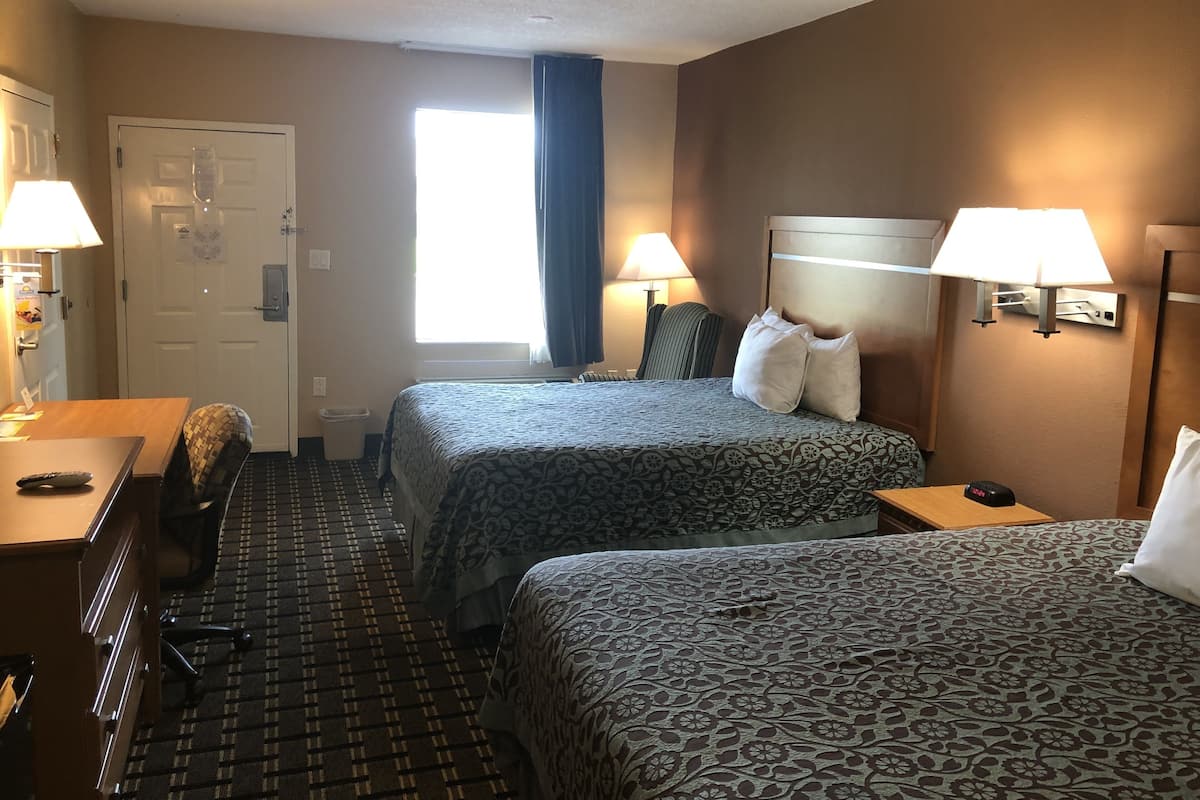 A hotel room with two beds, a desk, a chair, a trash bin, and a window with curtains.